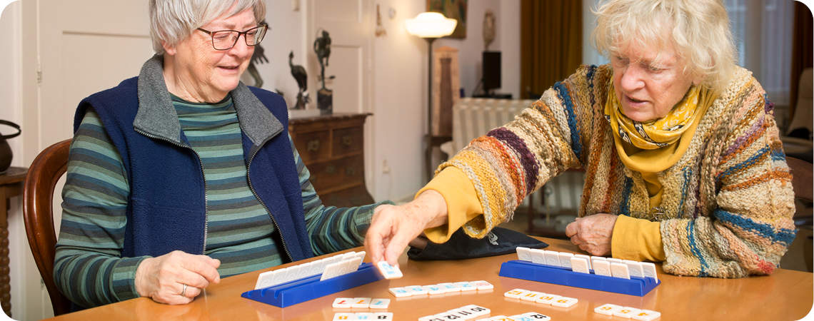 Op de foto twee bejaarden die in de huiskamer van het verzorgingshuis een spelletje Rummikub spelen. Dankzij de XXL versie van Rummikub die extra grote onderdelen heeft kunnen zij ook weer moeiteloos genieten van dit gezelschapspel voor ouderen. Ook op deze foto is weer goed zichtbaar hoe goed het is voor ouderen om spelletjes te spelen, zowel sociaal als mentaal en cognitief stimuleert deze speelse activiteit.